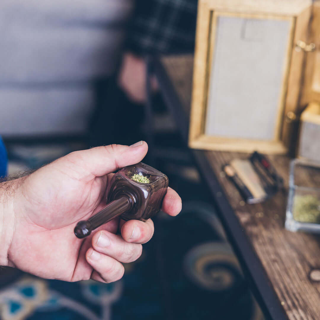 Hand holding cannabis pipe with Dexter Co setup in background”